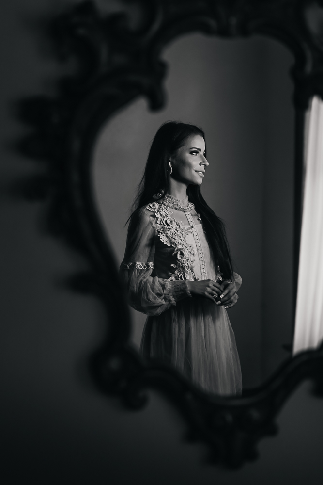 Black and white fine art portrait of a woman reflected in an ornate mirror while holding purple flowers.