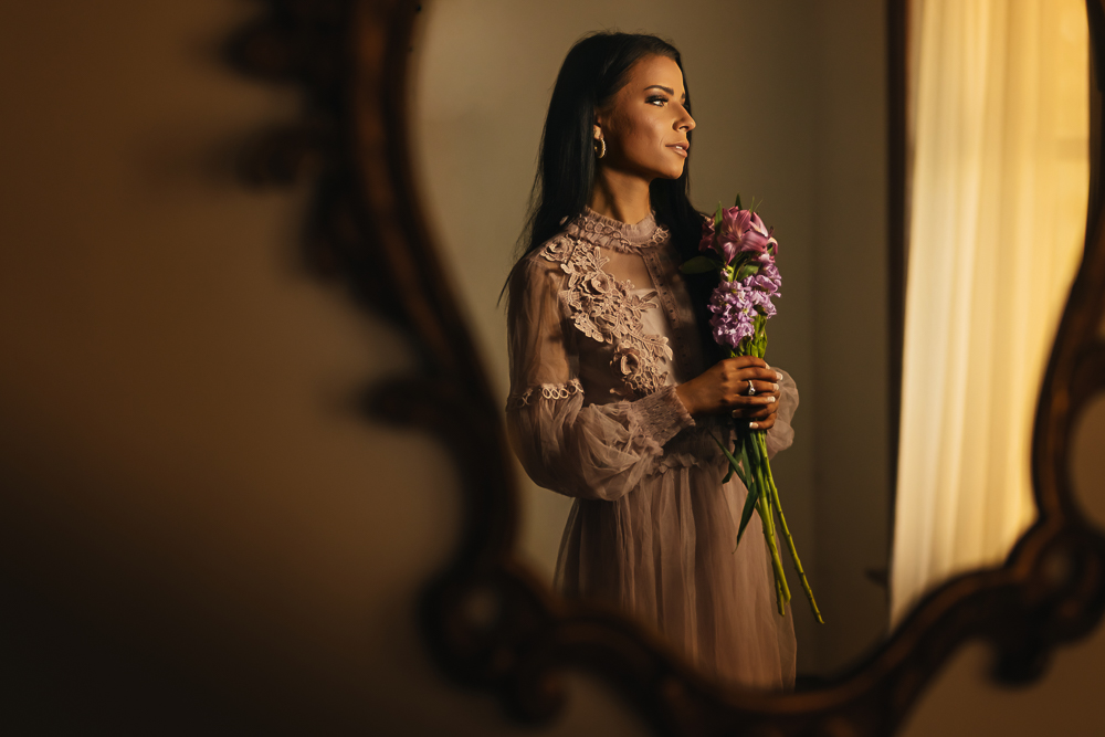 Woman with long dark hair holding a bouquet while standing in the Grand Foyer with elegant natural light.
