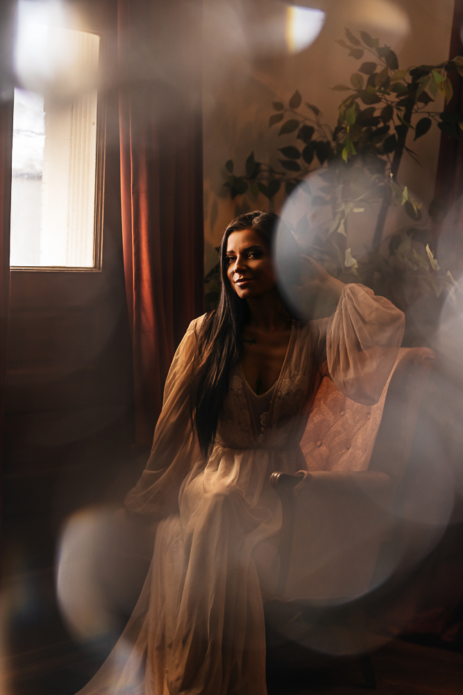 Fine art boudoir portrait of a brunette woman sitting beside a window in a sheer robe with soft natural light.