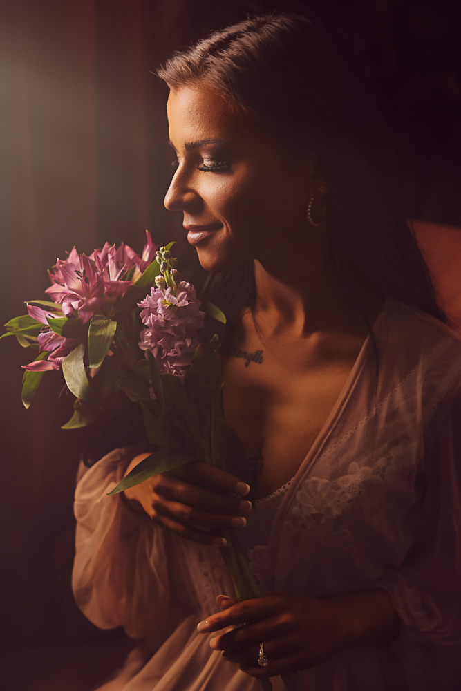 Side profile of a woman with long dark hair holding purple flowers in warm window light at Lafleur Boudoir.